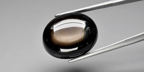 Black oval cab 6-ray star sapphire gemstone with smooth surface, held in silver tongs on a light gray background.
