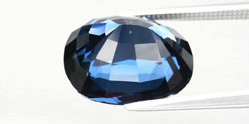 Large oval faceted blue spinel gemstone with black inclusions, held by a silver-tone pick against a white background.