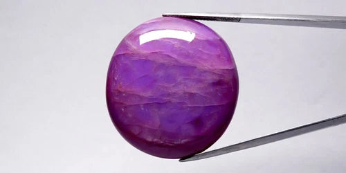 Unheated 124.62ct oval purple sapphire with 6 rays star pattern, held by silver-tone tweezers on white background.
